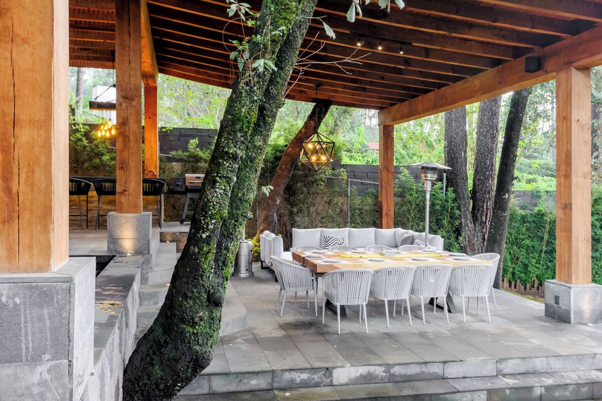 An outdoor patio with a wooden pergola, stone flooring, and a round table with white chairs.