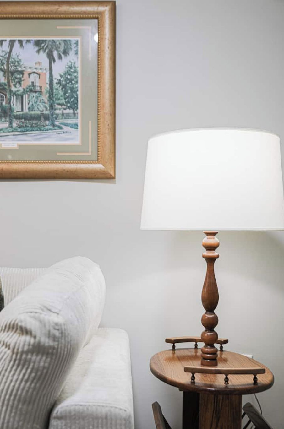 Een kamer met een witte bank, een houten tafel met een witte lampenkap en een lijst met een afbeelding aan de muur.