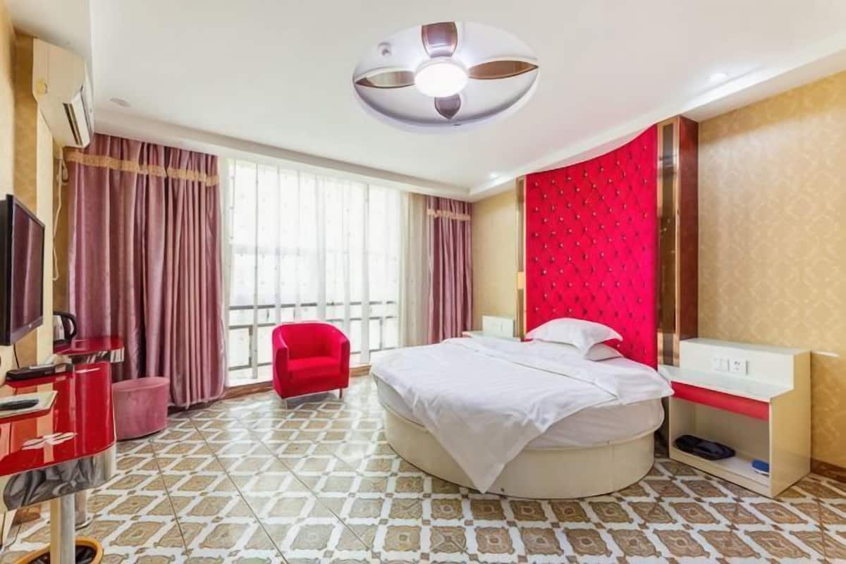 A hotel room with a round bed, a red chair, a TV, and a large window with curtains.