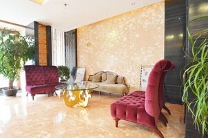 Un moderno vestíbulo con un sillón rojo acolchado, una mesa de centro de vidrio con base dorada, un sofá beige y una planta grande en maceta.