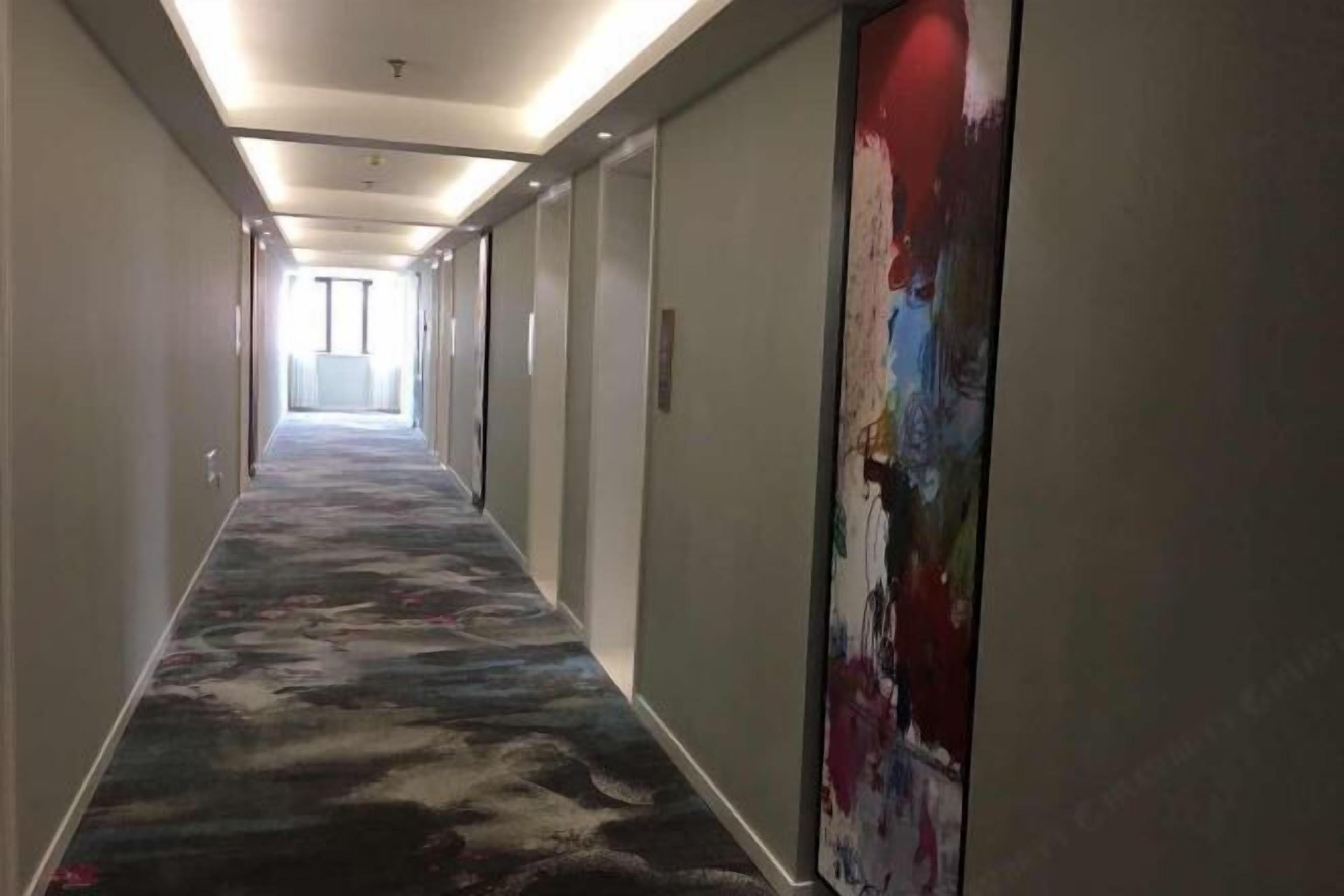 A hotel hallway with a carpeted floor, a large abstract painting on the right wall, and a door at the end.