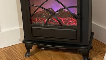 A black wood-burning stove with a glass door, placed on a wooden floor against a white wall.