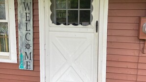 A white door with a decorative window above it, a 'WELCOME' sign to the left, and a doormat at the entrance. - Charming 3-bedroom cottage in Hampden with AC, WiFi (Hampden)