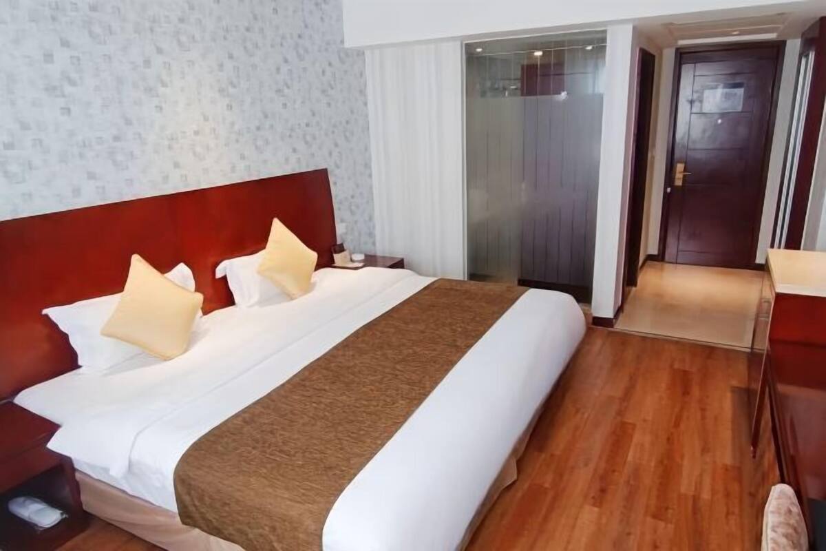 A hotel room with a large bed, a wooden floor, a red headboard, and a glass door leading to a bathroom.