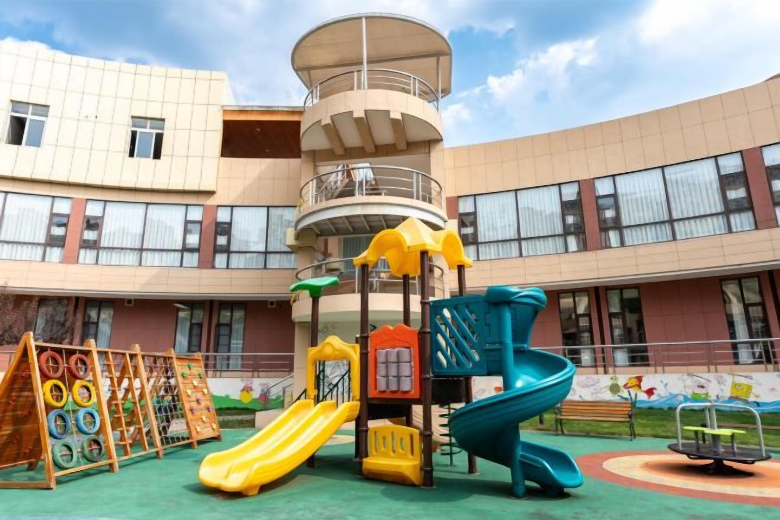 A playground with slides and climbing structures in front of a building with multiple windows and a balcony.