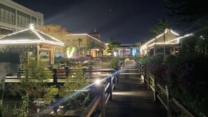 A nighttime outdoor area with wooden walkways, illuminated by overhead lights, surrounded by greenery and buildings. - Chengdu Star Flower Garden Hotel (University Games Sports Center) (Chengdu)