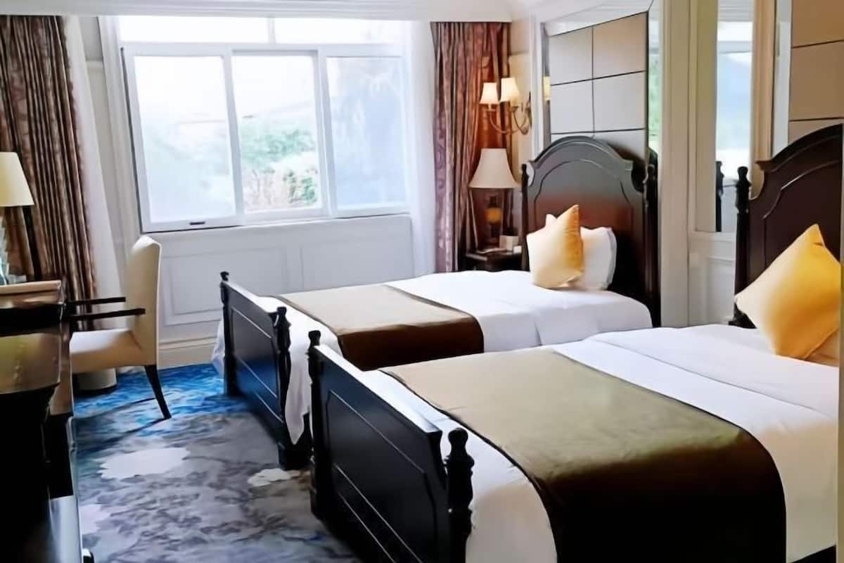A hotel room with two beds, a desk, a chair, a window, and a lamp.
