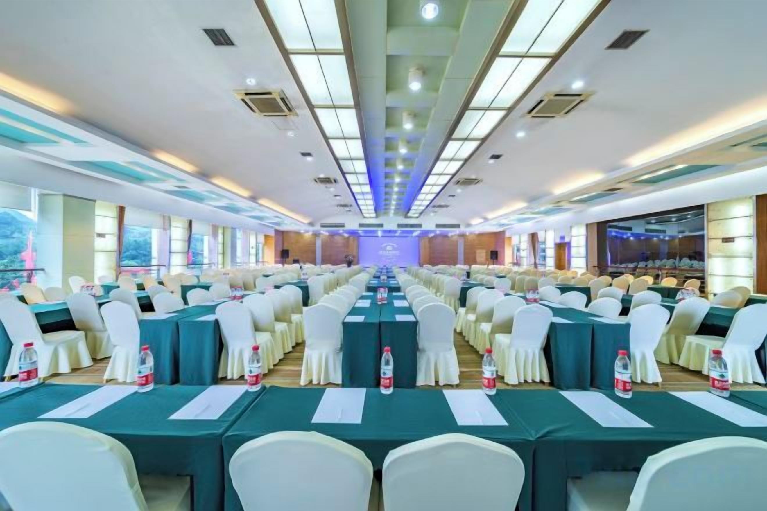 Un salón de conferencias con filas de mesas y sillas, un escenario al frente y un ventanal grande en un lateral.