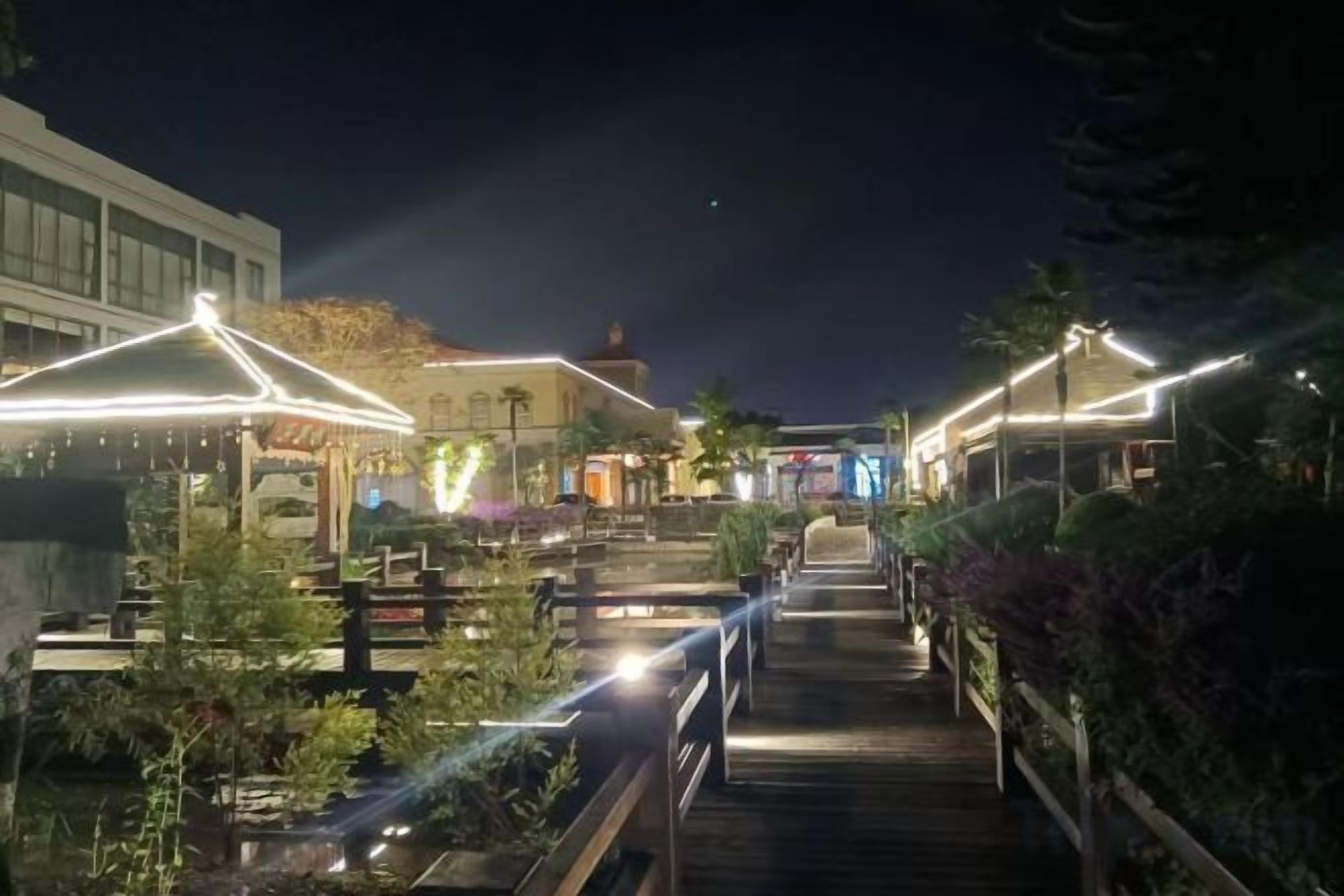 Un espacio al aire libre iluminado por luces colgantes, con un sendero de madera que conduce a un edificio con techo en forma de cúpula.