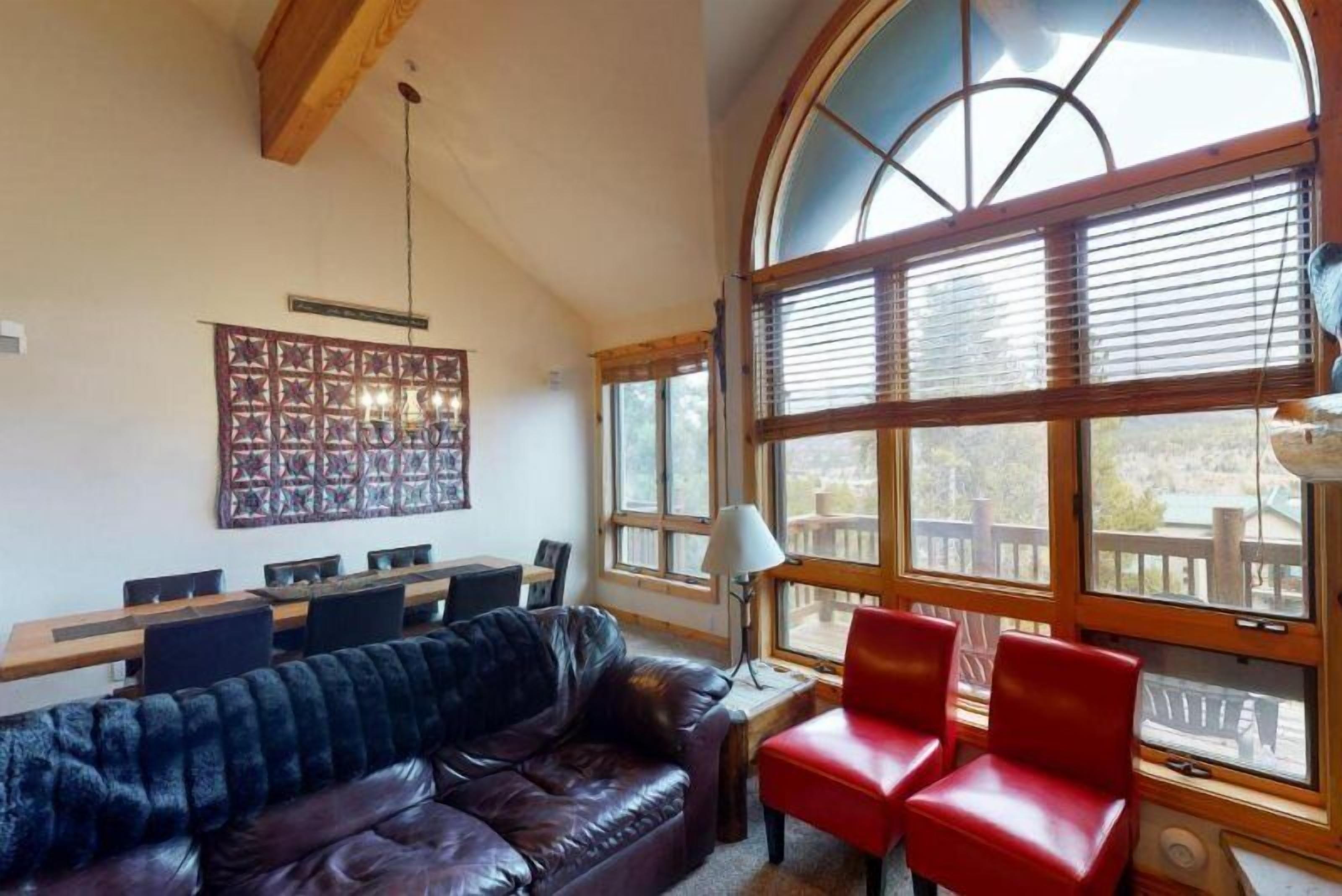 Una habitación acogedora con un sofá de cuero, sillas rojas, una mesa de comedor de madera y un gran ventanal con persianas.