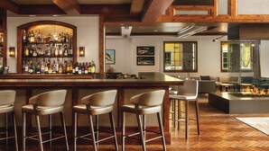 A bar area with a wooden bar counter, multiple stools, and a variety of bottles. There are framed pictures on the wall and a large window with a view of the outdoors. - The Ritz-Carlton Club®, Vail, Colorado. 2 Bedroom + DEN. 8PAX (Vail)