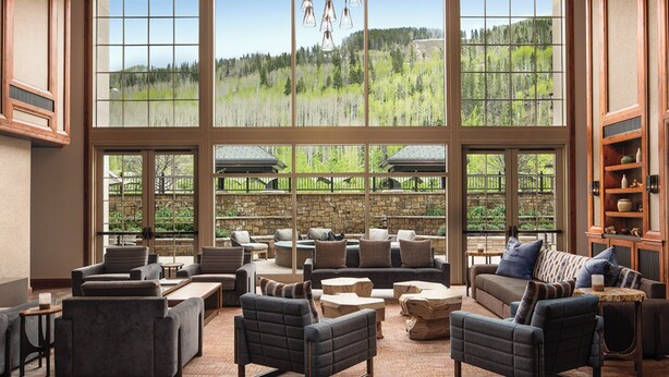 A spacious lounge area with multiple seating options, large windows offering a view of a forested hillside, and a stone wall in the background. - The Ritz-Carlton Club®, Vail, Colorado. 2 Bedroom + DEN. 8PAX (Vail)