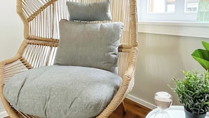 A wicker chair with grey cushions, a small table with a potted plant, and a window with a view of buildings. - Secretapartment: Zentral und Gemütlich, mit Großem Balkon (Warburg)