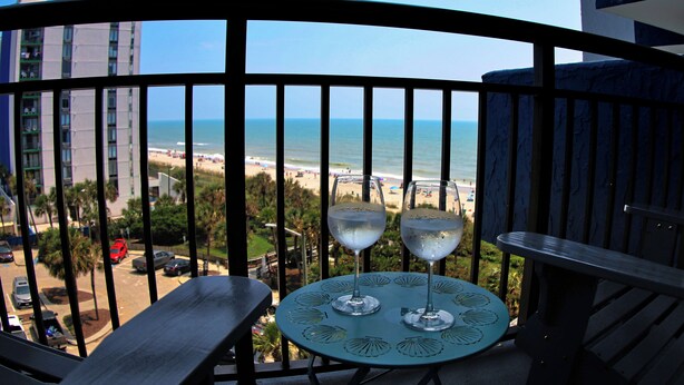 Un balcón con una mesa y dos vasos de agua, con vistas a una playa y al océano.