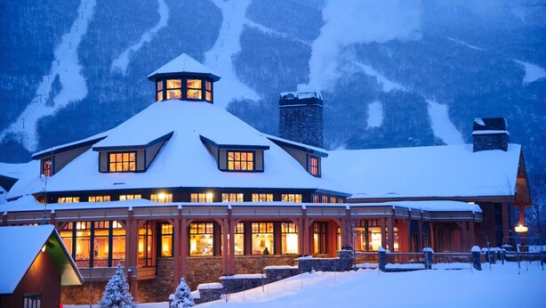 Un chalet enneigé, doté d’une tour imposante et de nombreuses fenêtres, se dressant sur un fond de montagnes.