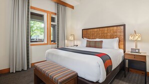 Une chambre d’hôtel avec un grand lit, une tête de lit en bois, une table de chevet avec une lampe, un banc et une fenêtre avec des rideaux.
