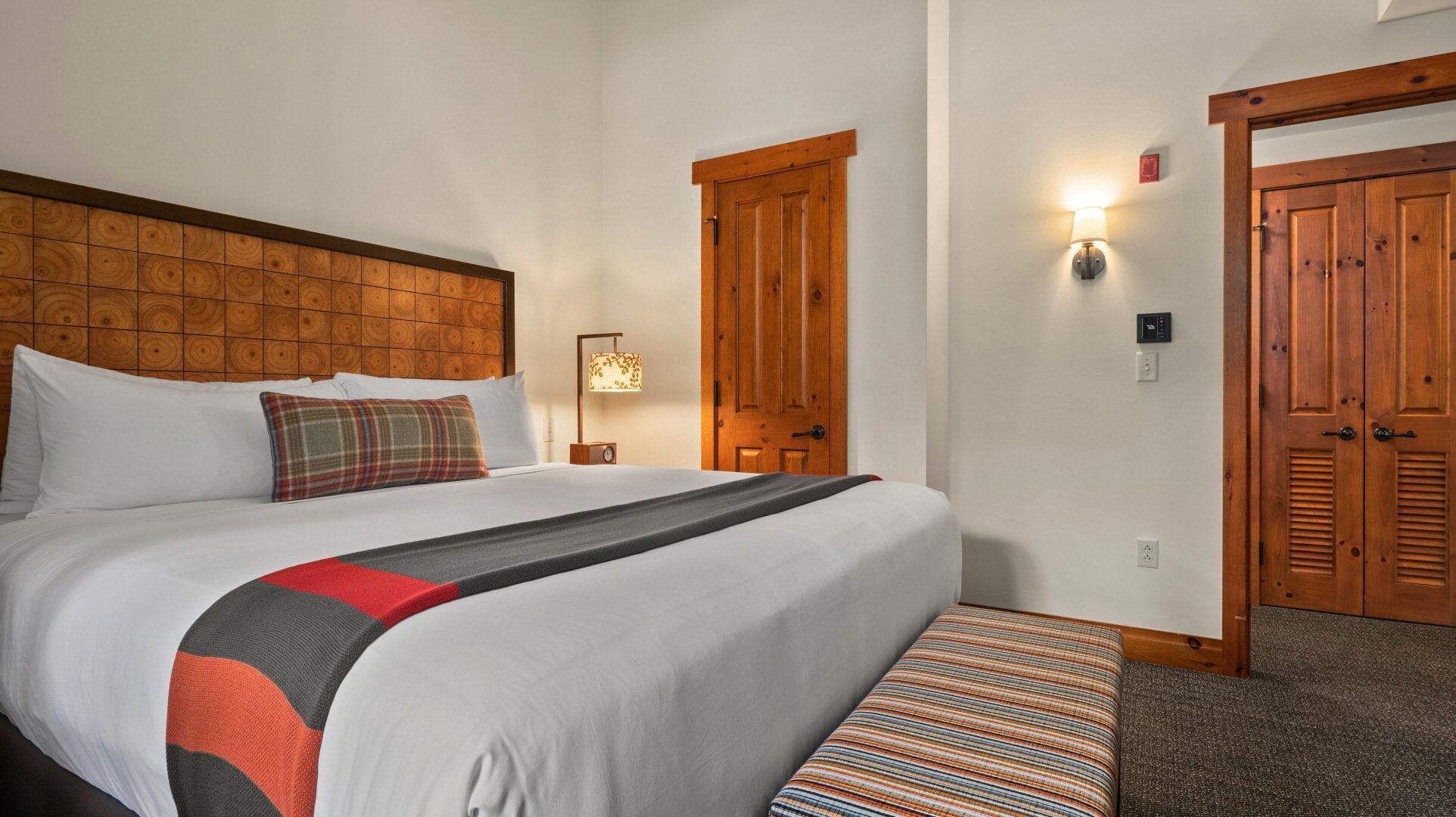 A hotel room with a large bed, a headboard with a circular pattern, a bedside lamp, a bench, and two wooden doors.