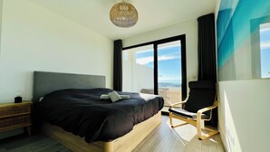 A modern bedroom with a large bed, a wooden nightstand, a chair, and a view of the city through large windows.