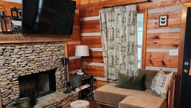 A cozy cabin-style room with a stone fireplace, wooden walls, a flat-screen TV, and a sofa with cushions.
