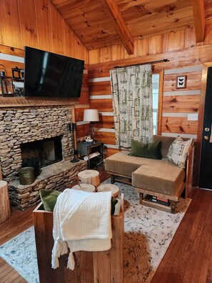 Une chambre confortable, aménagée dans le style d’un chalet, avec une cheminée en pierre, des murs en bois, un téléviseur à écran plat et un canapé avec des coussins.