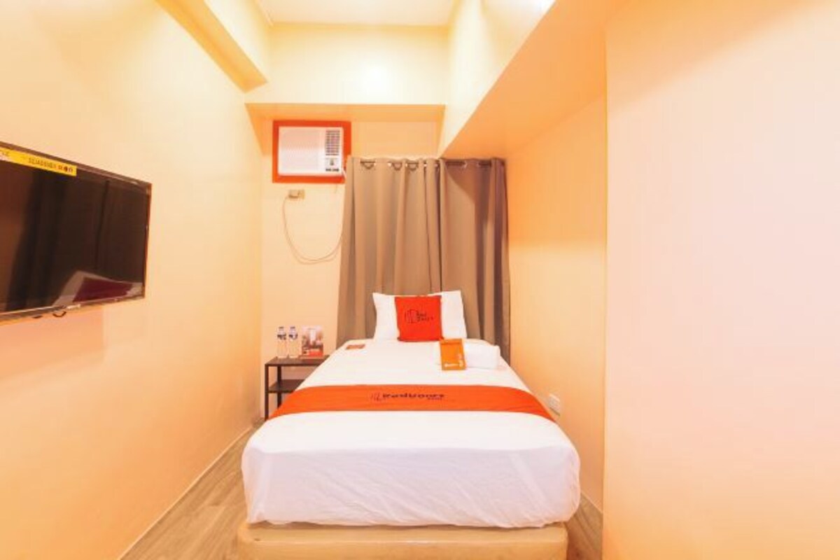 Une chambre d’hôtel avec un lit, un téléviseur fixé au mur, un rideau et une petite table avec une lampe.
