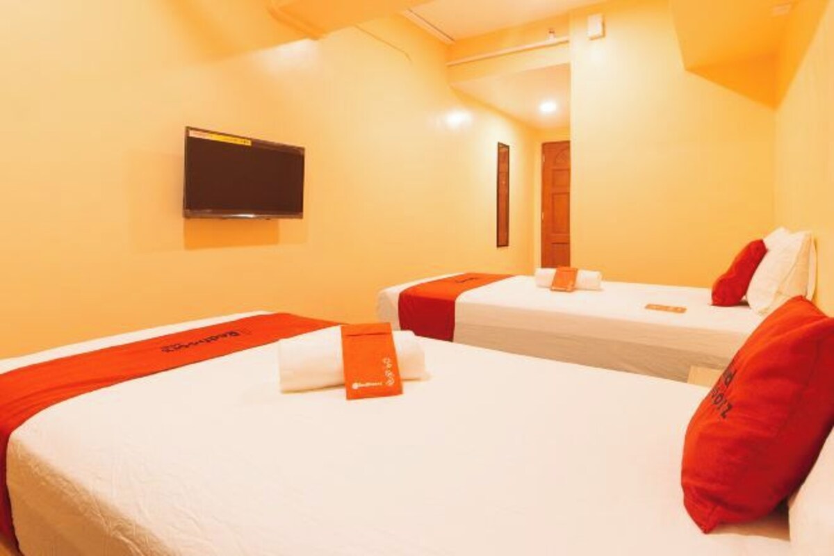 Une chambre d’hôtel avec deux lits, chacun agrémenté de draps blancs et de touches de couleur rouge, un téléviseur à écran plat fixé au mur et une porte en bois.