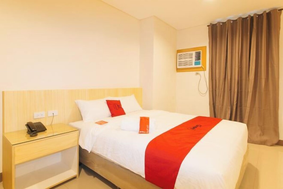 A hotel room with a bed, a nightstand with a phone, a wall-mounted air conditioner, and a window with curtains.