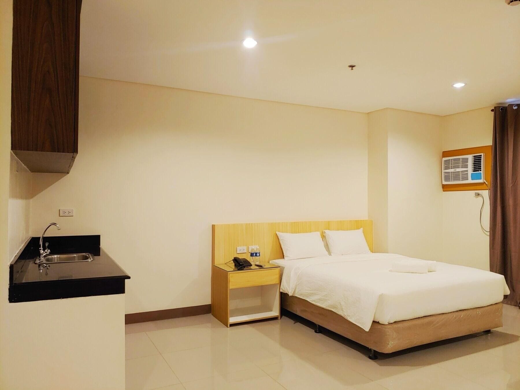 Una habitación de hotel con una cama, una mesita de noche, un escritorio pequeño con un teléfono, un lavamanos y un equipo de aire acondicionado.