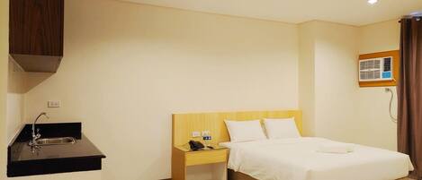 A hotel room with a bed, a nightstand, a small desk with a phone, a sink, and an air conditioning unit.