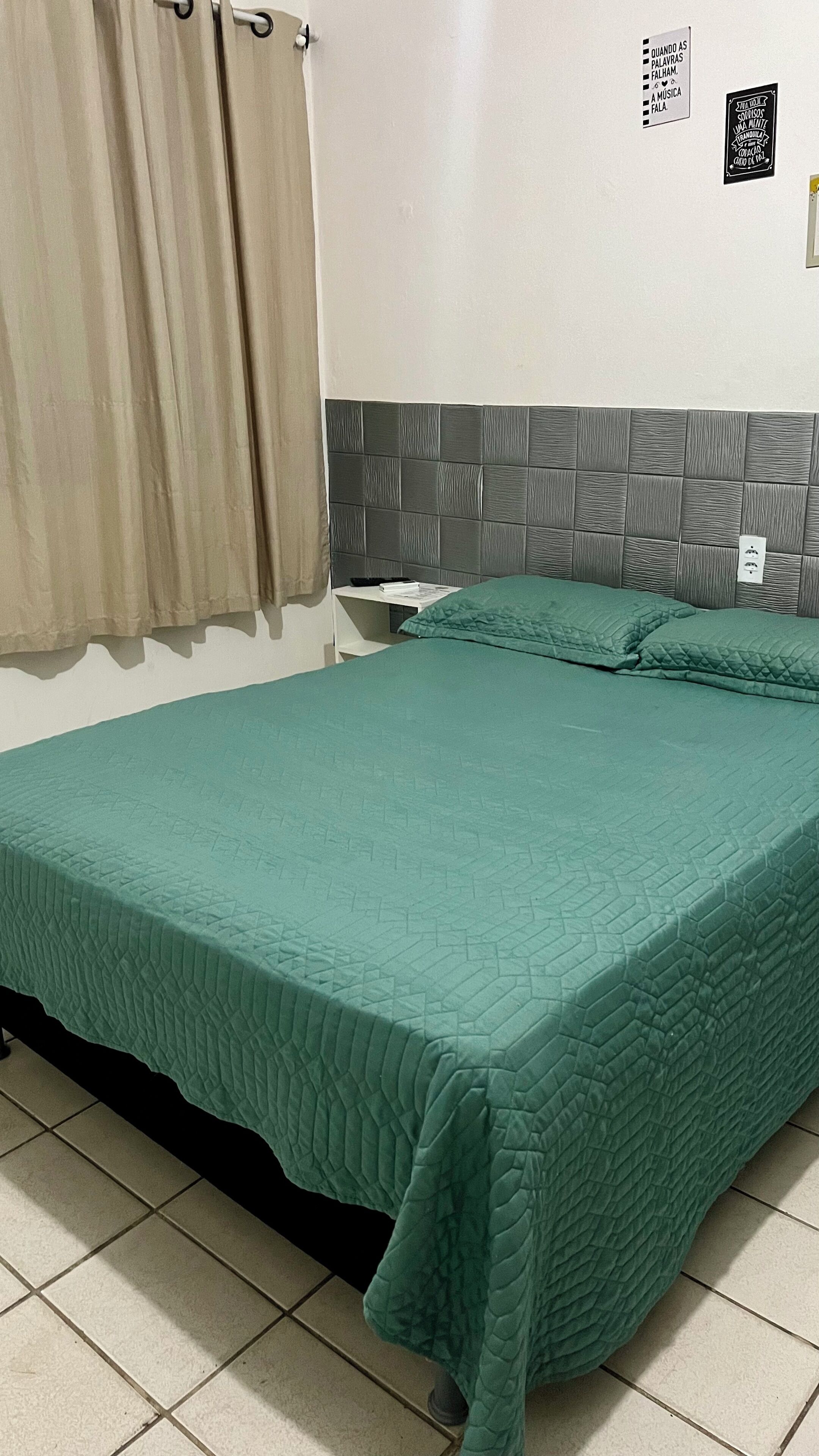 A bedroom with a green quilted bedspread, a grey headboard, and a curtain on the window.