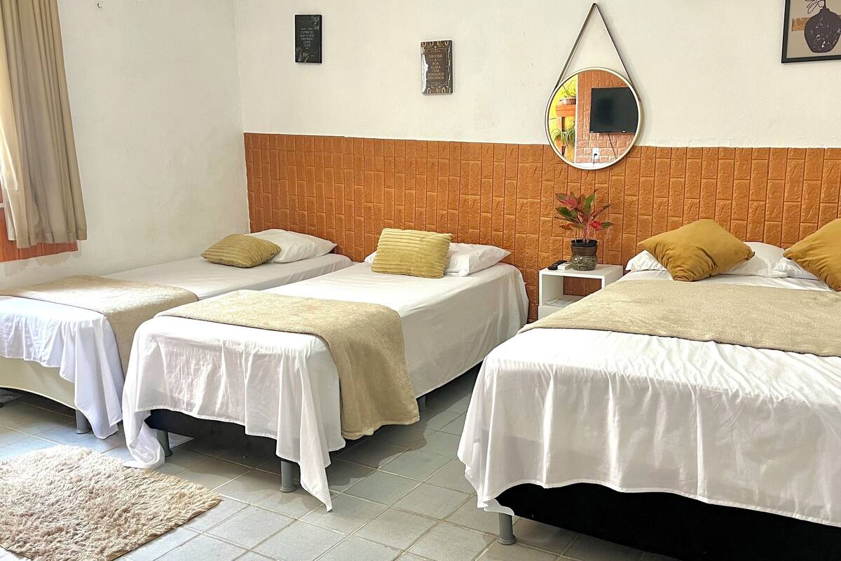 A hotel room with two single beds and one double bed, each with white bedding and decorative pillows. There is a small bedside table with a lamp, a mirror on the wall, and a framed picture. The room has a tiled floor and a window with curtains.