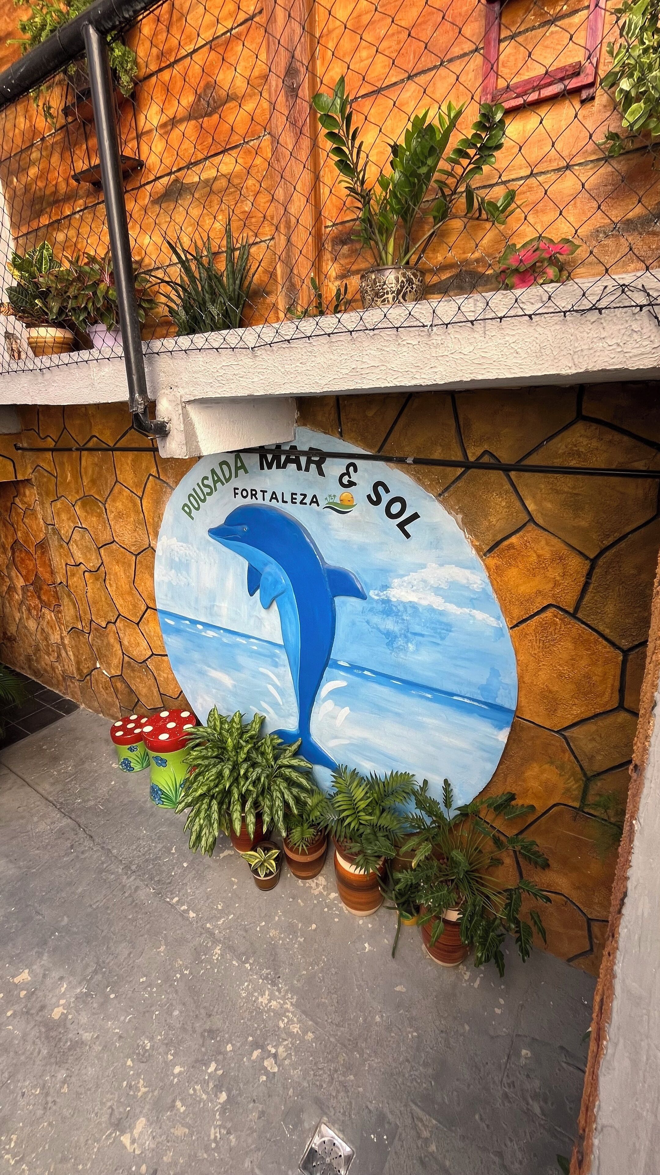 Paredes com mural de um golfinho e os textos "POUSADA MAR & SOL" e "FORTALEZA". Vasos com plantas no chão.