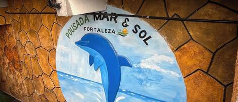 Paredes com mural de um golfinho e os textos "POUSADA MAR & SOL" e "FORTALEZA". Vasos com plantas no chão.
