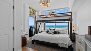 A bedroom with a bunk bed, a desk, a chair, a lamp, and a view of the ocean.