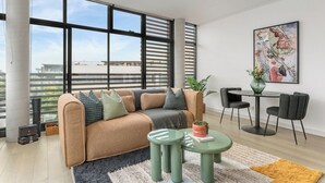 Una sala de estar moderna con sofá, mesa de centro y sillas, ventanas amplias con persianas, un cuadro en la pared y vistas a edificios.