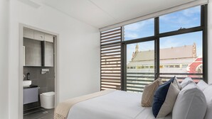 Un dormitorio moderno con una cama grande, un armario empotrado, un baño y vistas a un edificio a través de amplios ventanales.