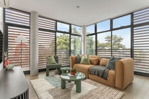 A modern living room with a sofa, armchairs, a coffee table, and large windows offering a view of trees. - King Street Apartments by Urban Rest (Newtown)