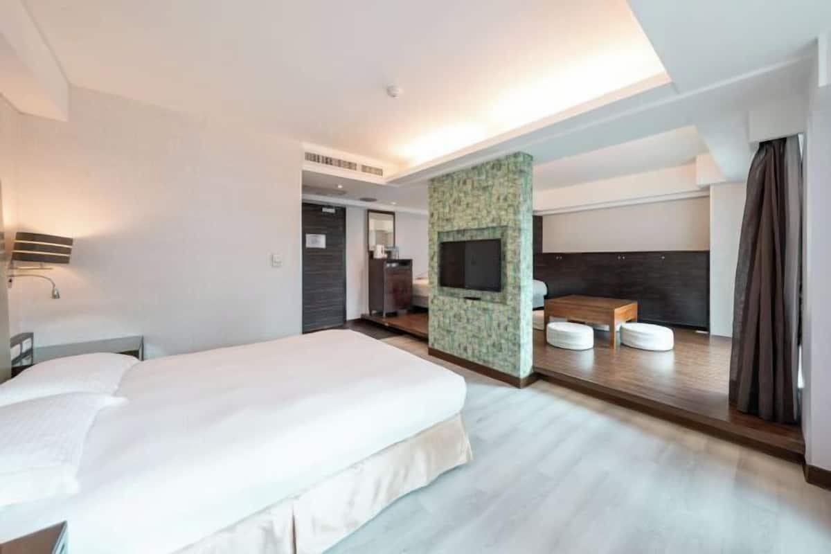 A modern hotel room with a large bed, a seating area with cushions, a television, and a fireplace.