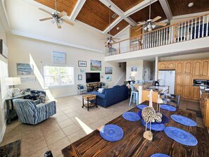 A spacious living area with a high ceiling, a dining table with blue placemats, a kitchen with wooden cabinets, and a balcony with a railing. - Beautiful Home in Gulf Shores with Private Pool, Sleeps 12 (Gulf Shores)