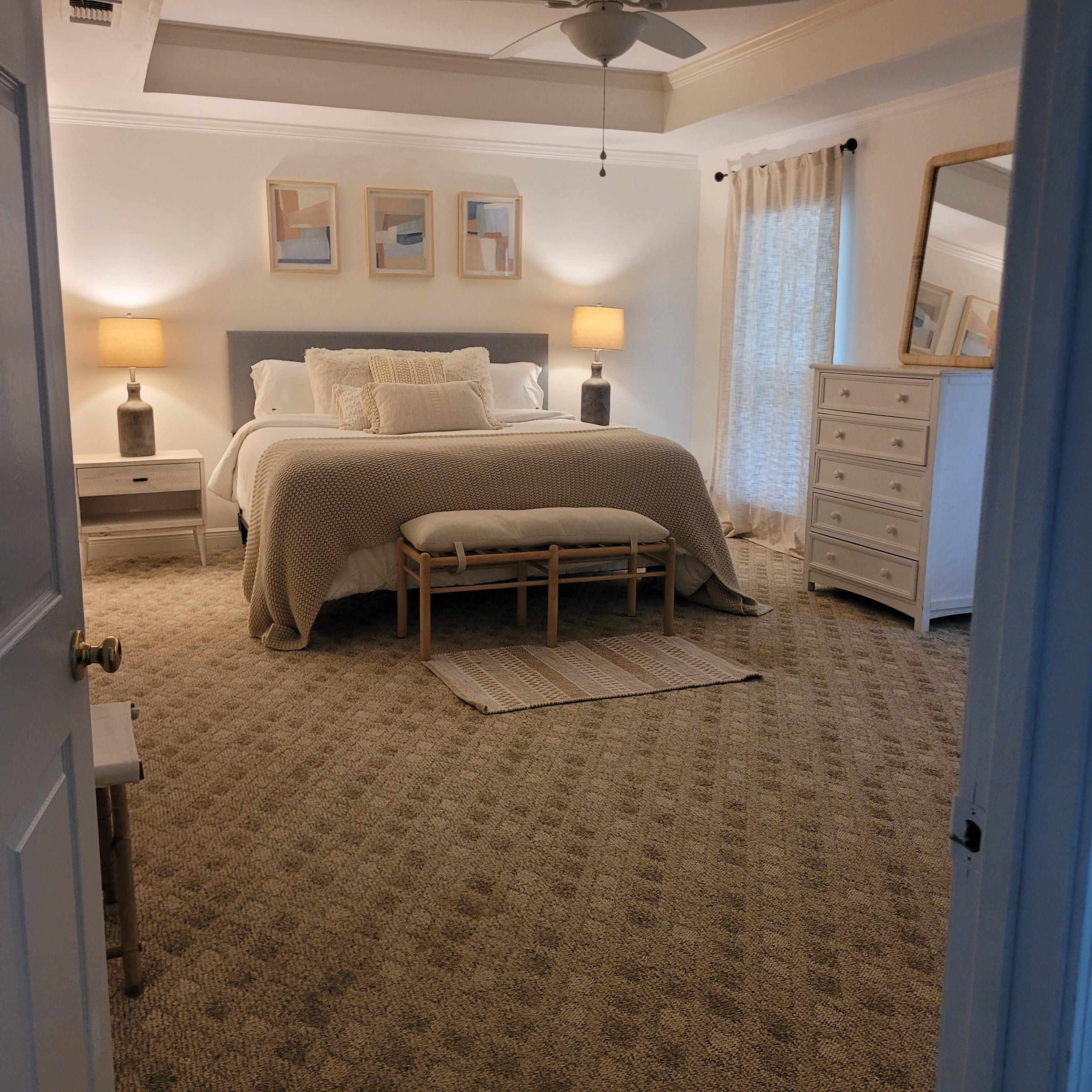 Une chambre à coucher avec un grand lit, deux tables de chevet avec des lampes, un banc, une commode et un ventilateur de plafond.
