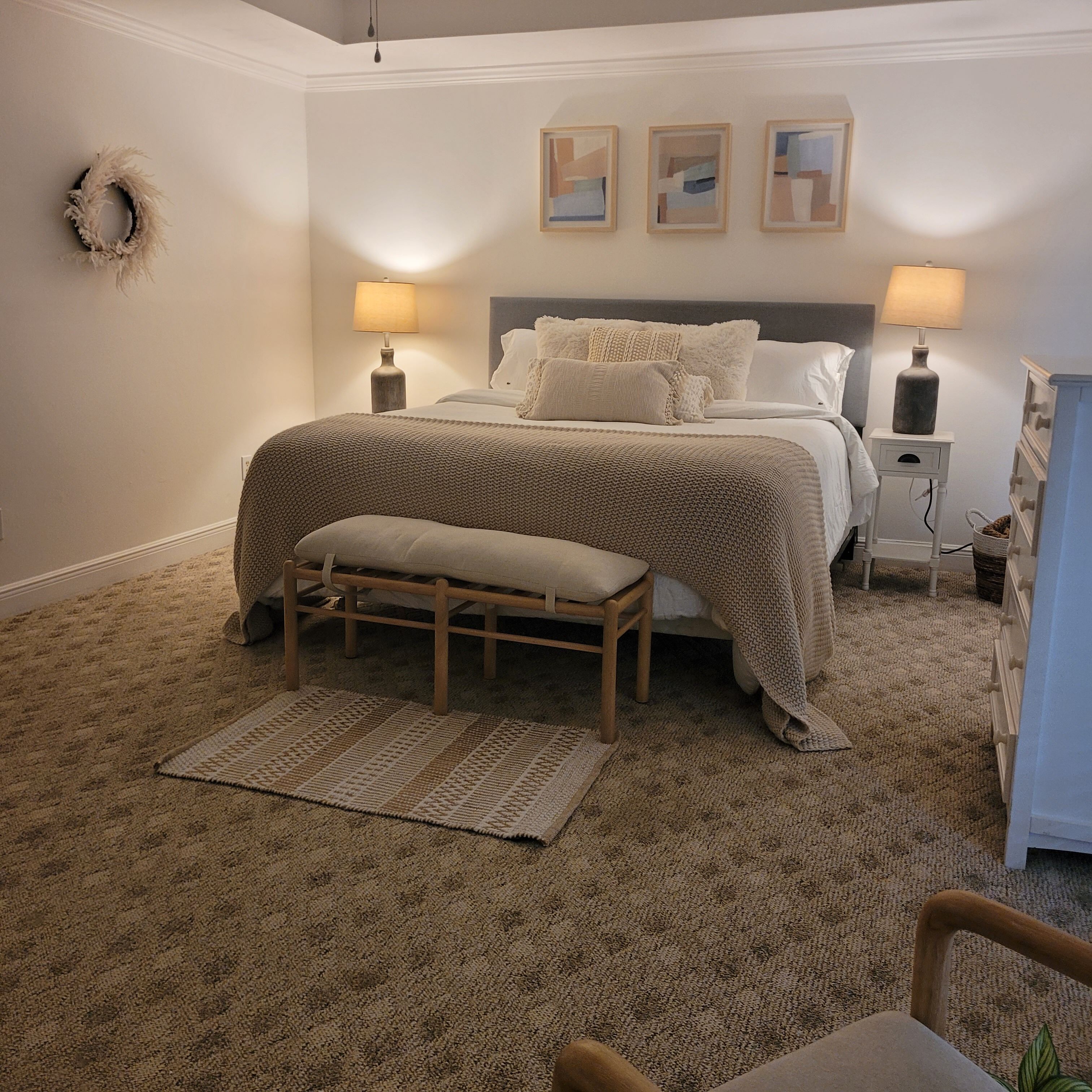 Une chambre à coucher avec un grand lit, deux lampes de chevet, un banc, un tapis et trois tableaux encadrés accrochés au mur.