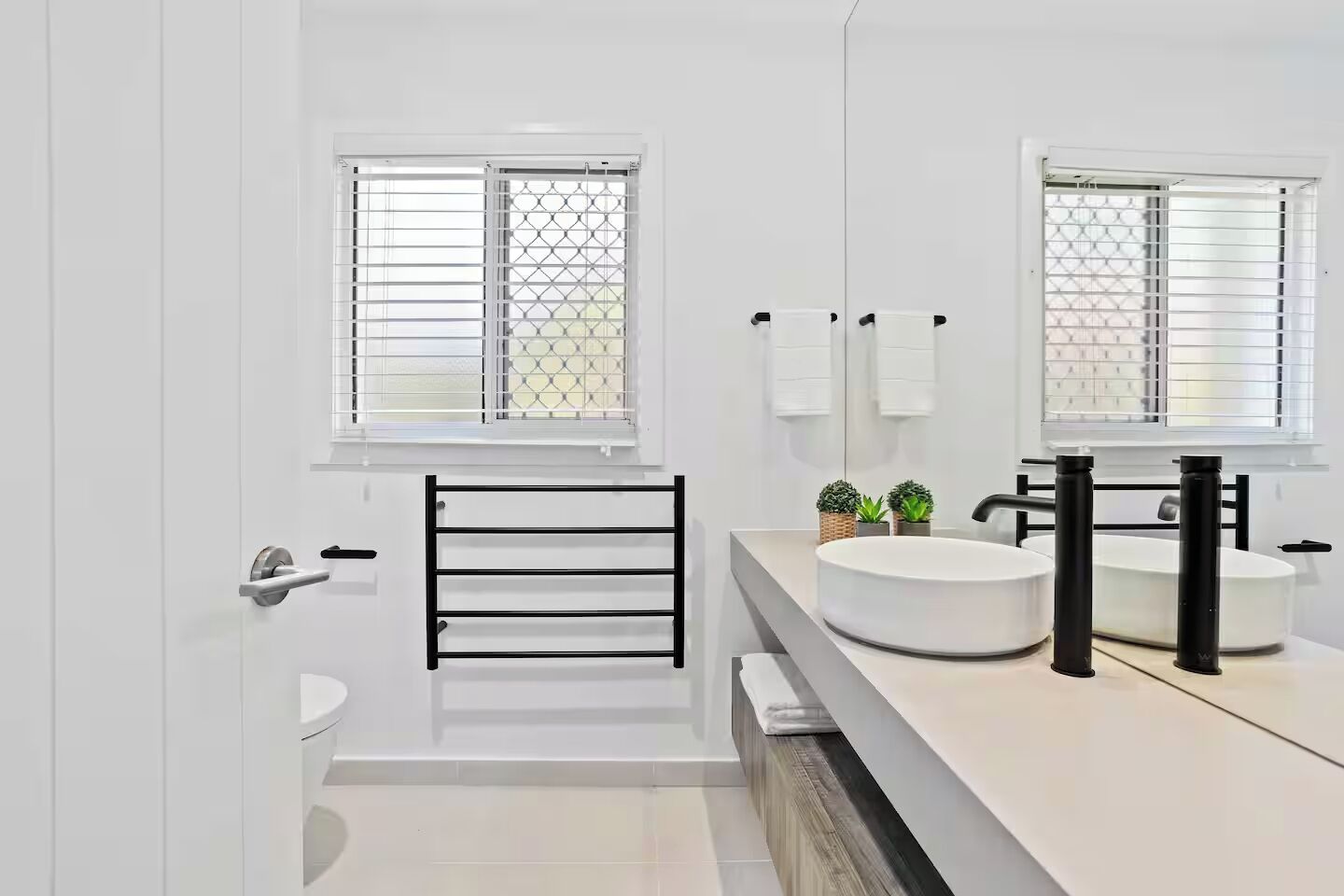 A modern bathroom with a white sink, two towel racks, a toilet, and a window with blinds.