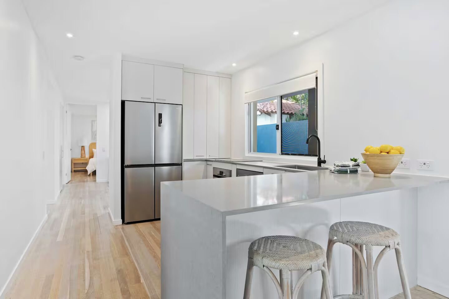 A modern kitchen with white cabinetry, a stainless steel refrigerator, a large island with bar stools, and a window with a view of a blue building.