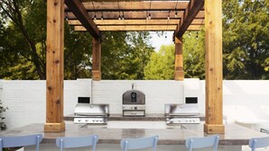 An outdoor barbecue area with a grill, a pizza oven, and two gas grills, surrounded by a wooden pergola and white brick walls. - 2xKing-Beds, Birkdale-Plaza-Vista, Shop-Eat-Play (Huntersville)