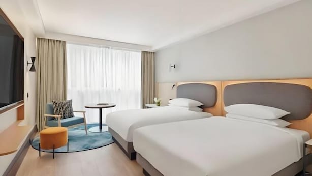 Ein Hotelzimmer mit zwei Betten, einem kleinen Tisch, einem Stuhl und einem großen Fenster mit Vorhängen.
