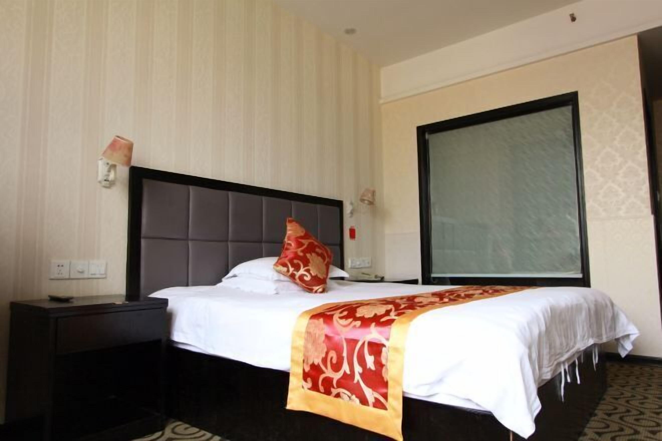 A hotel room with a bed, a nightstand, a wall-mounted lamp, and a large window.
