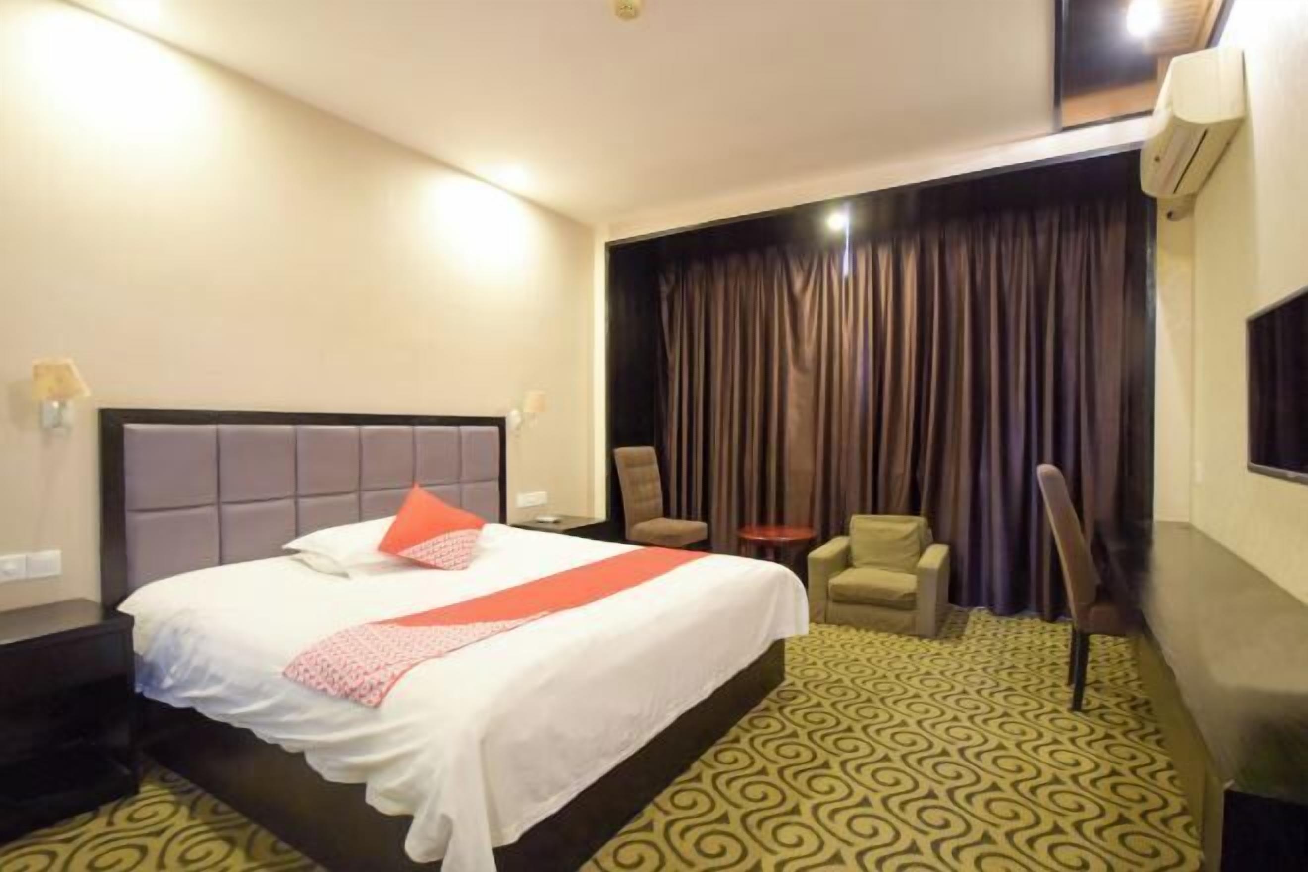 Quarto de hotel com uma cama grande, uma cadeira, uma mesinha e uma janela com cortinas.