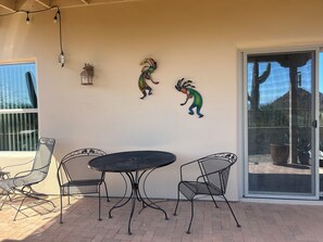 Une terrasse avec une table ronde et quatre chaises, une décoration murale représentant un paon, et une porte-fenêtre coulissante donnant sur une vue d’un cactus.
