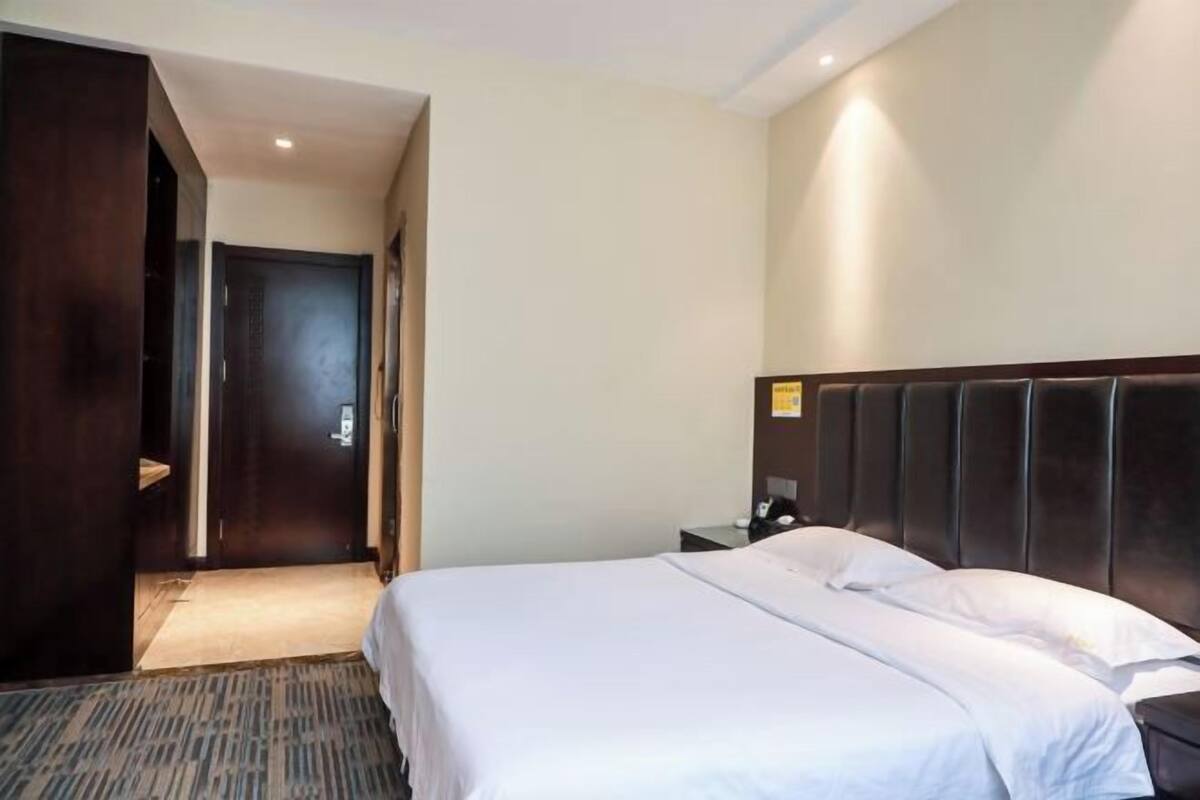 Une chambre d’hôtel avec un grand lit, une tête de lit sombre, une table de chevet et une porte donnant sur un couloir.