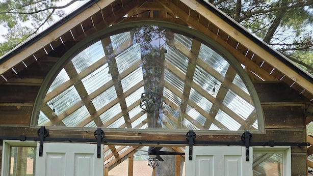 Une structure en bois surmontée d’un grand toit en verre en forme d’arche, avec un banc et des plantes en pot.